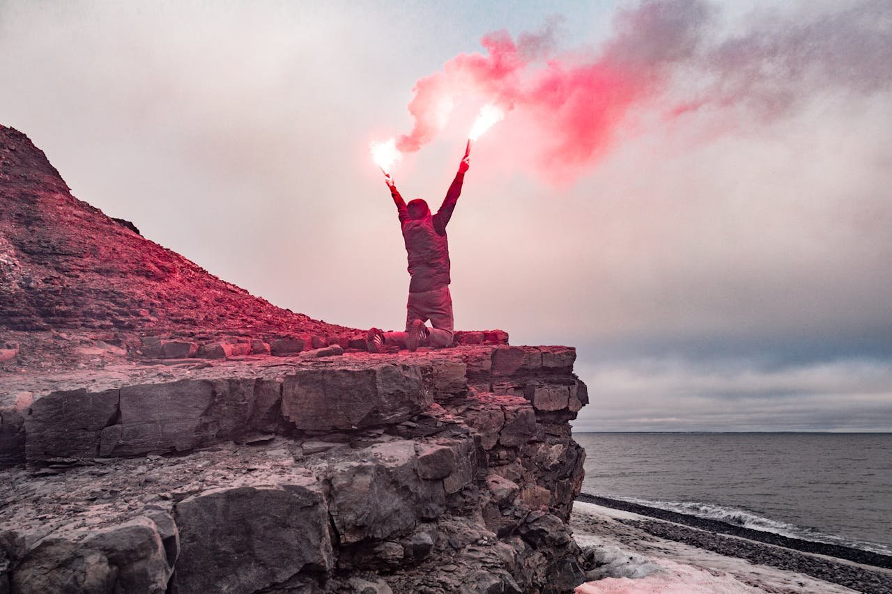 Dramatic scene of a man with flares signaling for help on a rocky cliff by the ocean.